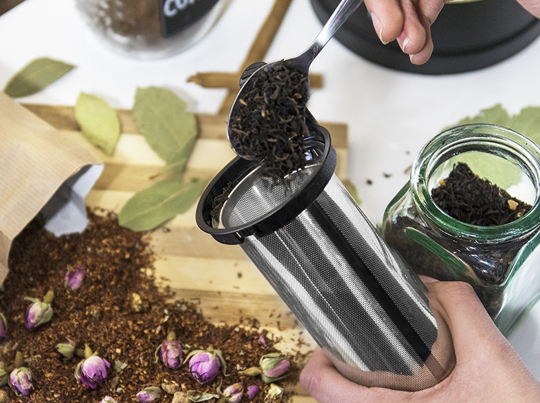 Una cuchara vierte hojas de té en un infusor de té plateado. Hay frascos de té e ingredientes.