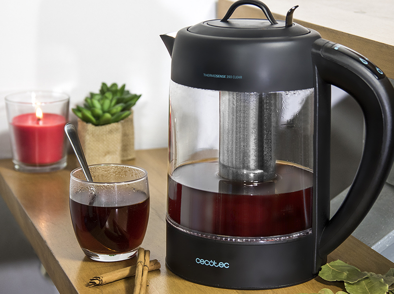 Hervidor eléctrico con té dentro. Vaso de té, velas y una planta sobre la superficie de madera.