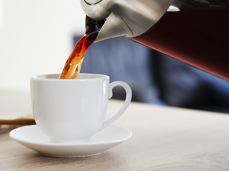 Café vertido desde una tetera plateada en una taza blanca sobre un platillo en una mesa de madera.