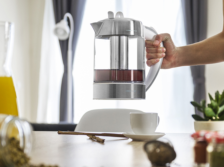 Una persona sosteniendo una tetera de vidrio con té dentro. Una taza y otros objetos están en la mesa.