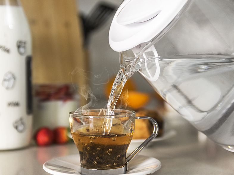 Vierte agua caliente de una jarra transparente en una taza de vidrio con té, sobre un platillo blanco.