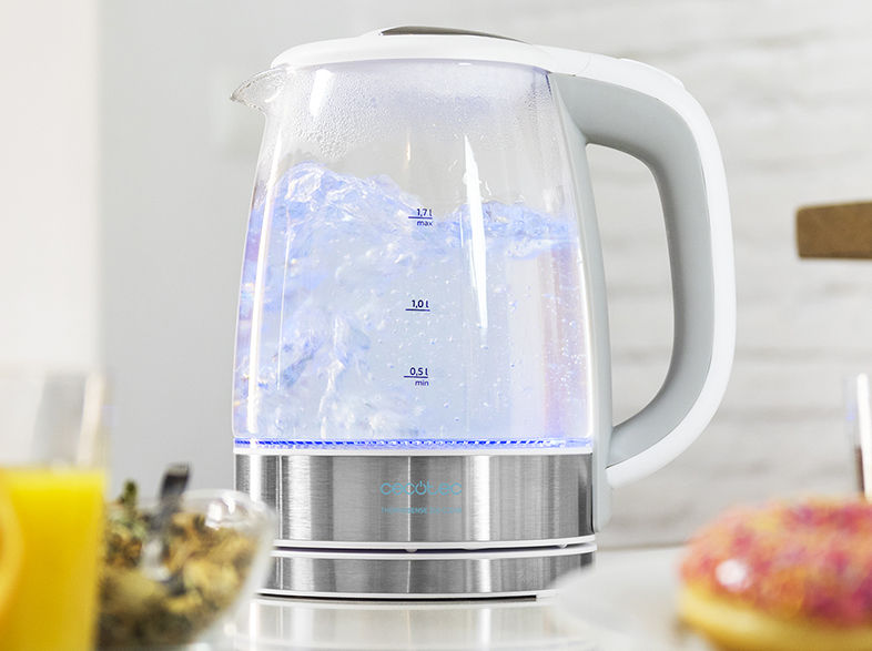 Hervidor de vidrio con agua hirviendo, luz azul y asa blanca. Comida y bebida en el fondo.