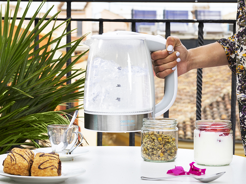 Una persona sosteniendo una tetera de vidrio con agua hirviendo, junto a pasteles y flores.