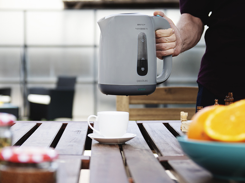 Un hombre sostiene una tetera gris; una taza y platillo están sobre una mesa.