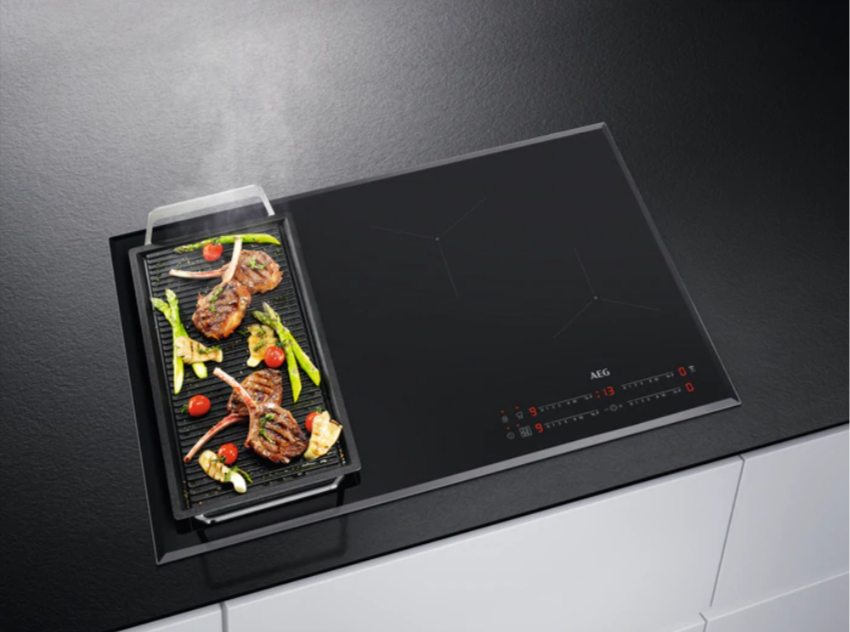 Zwarte inductiekookplaat met een grill aan de zijkant. Eten, inclusief groenten, ligt op de grill.