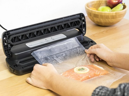 Persona sellando bolsa de salmón con selladora al vacío, mesa de madera, frutero.