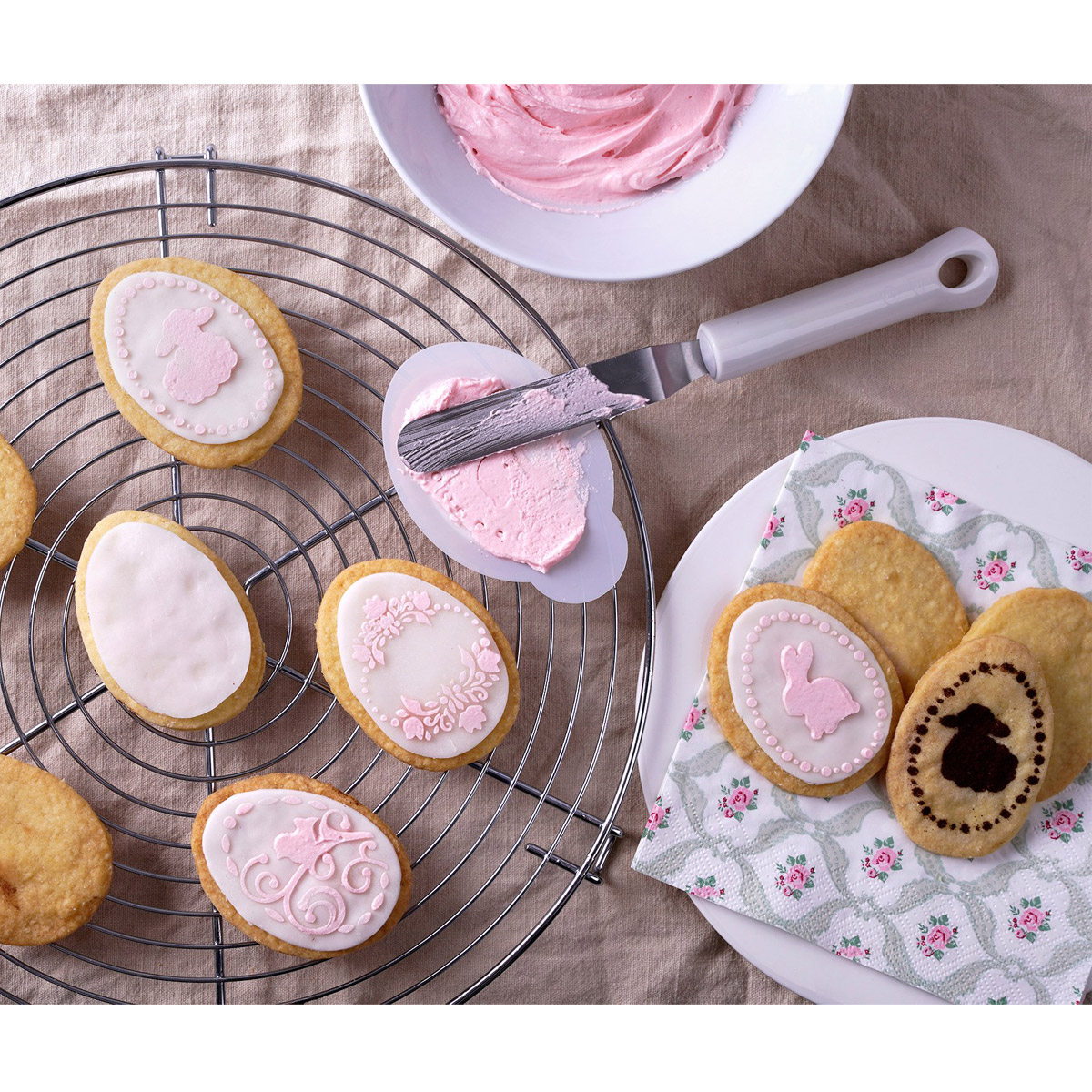 Kekse auf einem Drahtgestell. Rosa Zuckerguss, ein Spatel und ein weißer Teller mit dekorierten Keksen.