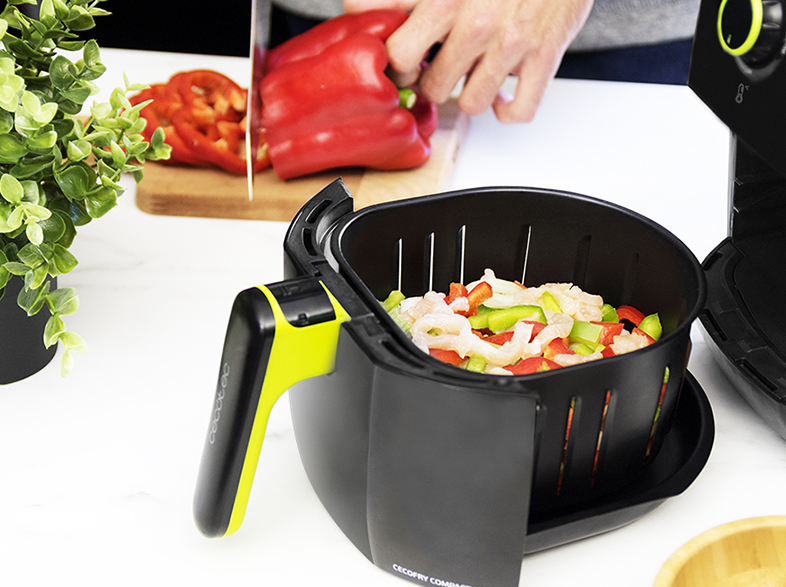 Una persona corta un pimiento rojo. Una cesta de freidora de aire contiene verduras picadas. Una planta está en el fondo.