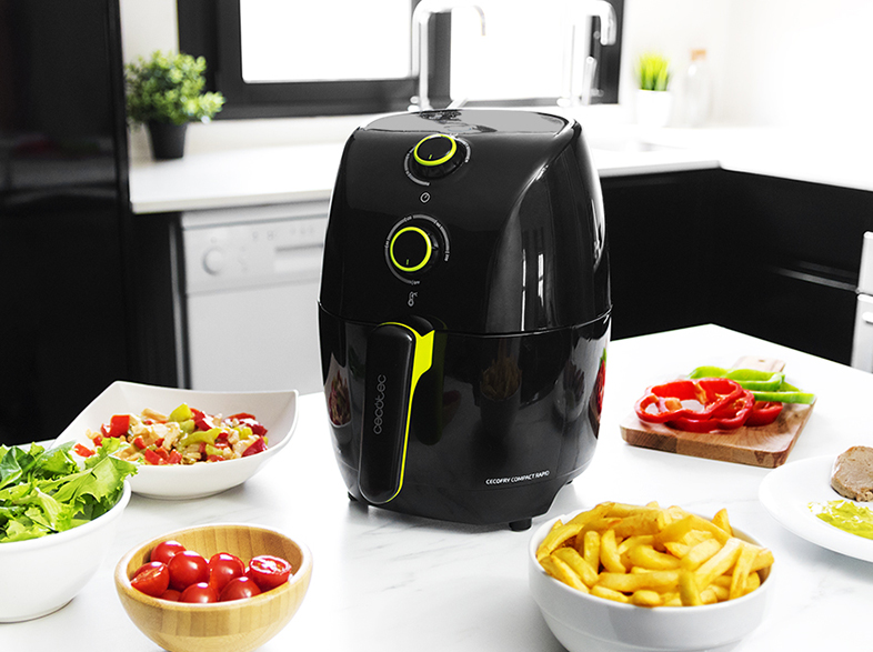 Freidora de aire negra con detalles amarillos. Cuencos con comida: ensalada, tomates, patatas fritas y pimientos. Fondo de cocina.