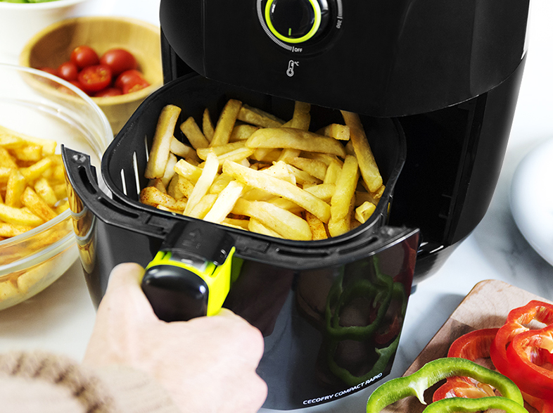 Freidora de aire llena de patatas fritas. Mano sosteniendo la cesta. Verduras y tomates al fondo.