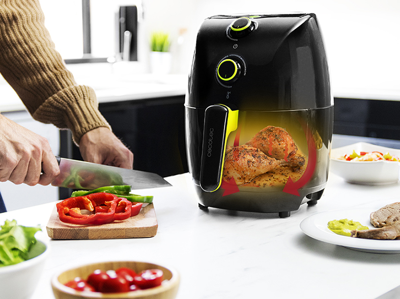 Una persona corta verduras. Una freidora de aire con pollo dentro está a la derecha. Platos de comida están a la derecha e izquierda.
