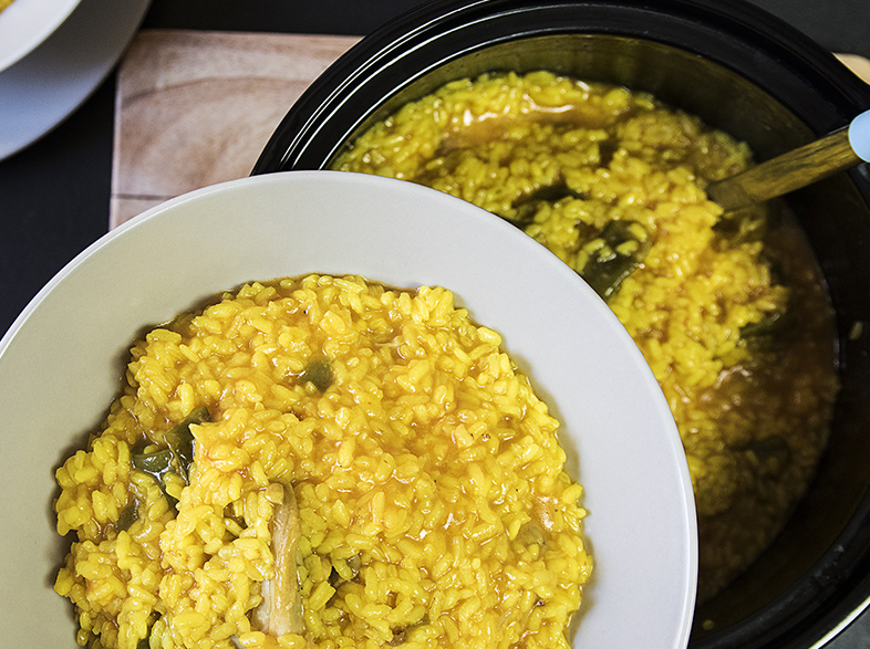 Un tazón de arroz amarillo con motas verdes, junto a una olla de arroz. Una cuchara de madera está en la olla. Hay platos blancos cerca.