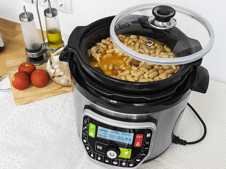 Una olla a presión con frijoles y carne dentro. Tomates, pan y botellas de aceite al lado.