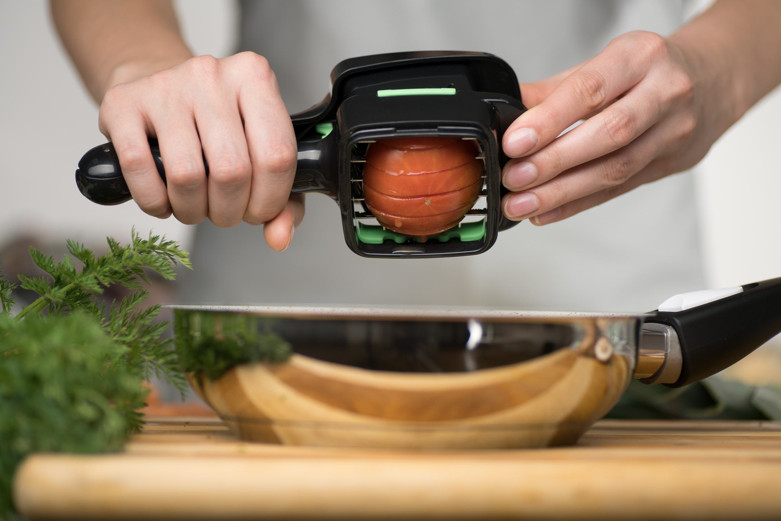 Hände schneiden eine Tomate mit einem Schneider über einer Pfanne.