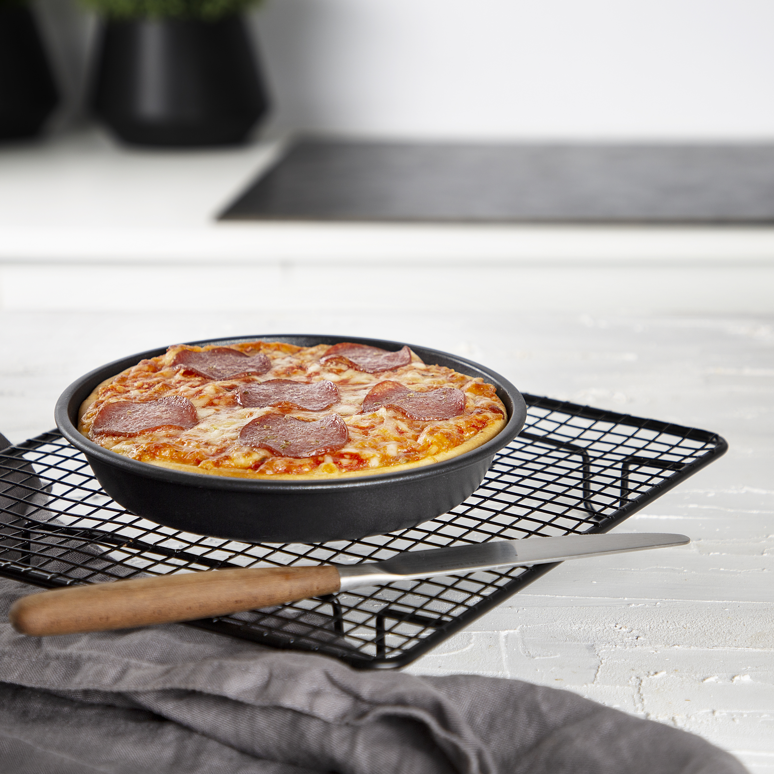 A pizza in a black pan on a wire rack, a knife, and a gray cloth on a white countertop.