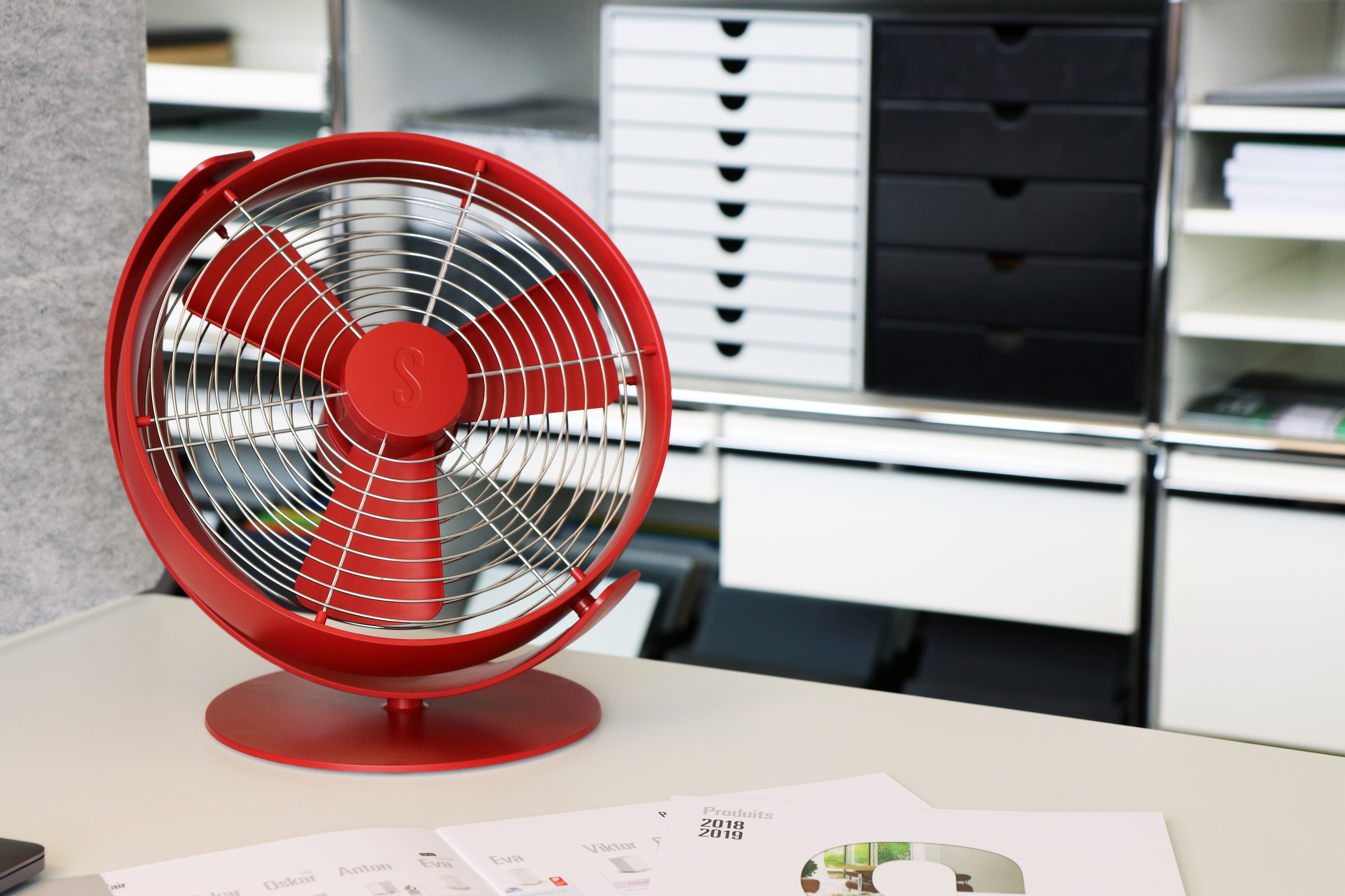 Ein roter Ventilator steht auf einer weißen Oberfläche. Dahinter sind Schubladen und Regale.