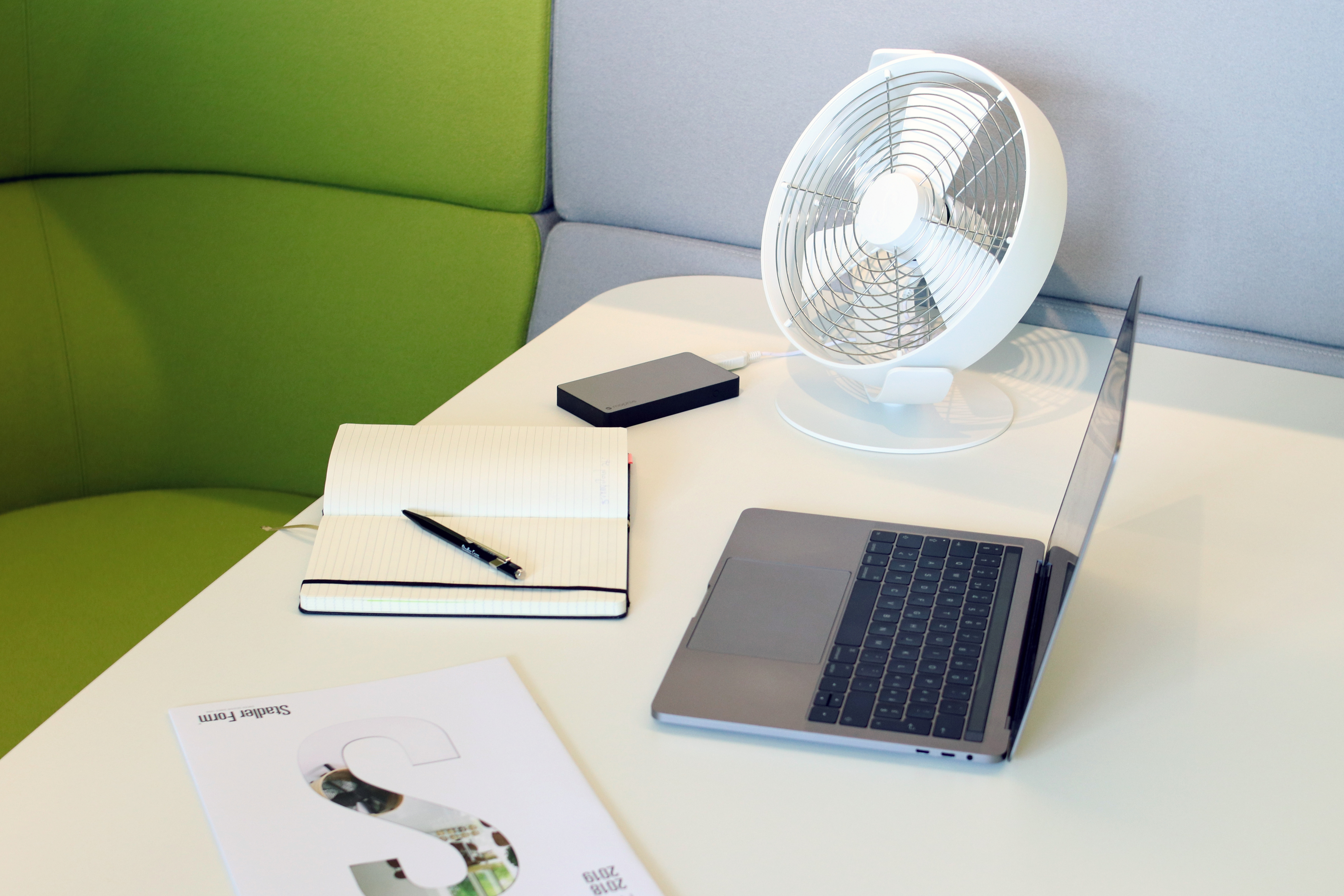 Ein Schreibtisch mit Laptop, Ventilator, Notizbuch, Stift und einem Dokument.