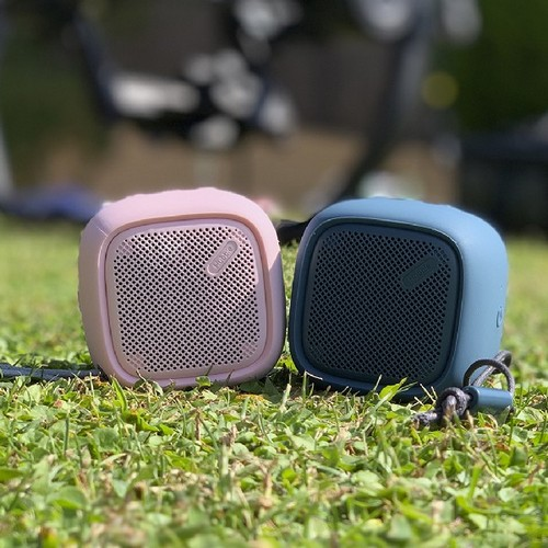 Dos altavoces pequeños y cuadrados, uno rosa y otro azul, sobre hierba verde. Un día soleado.
