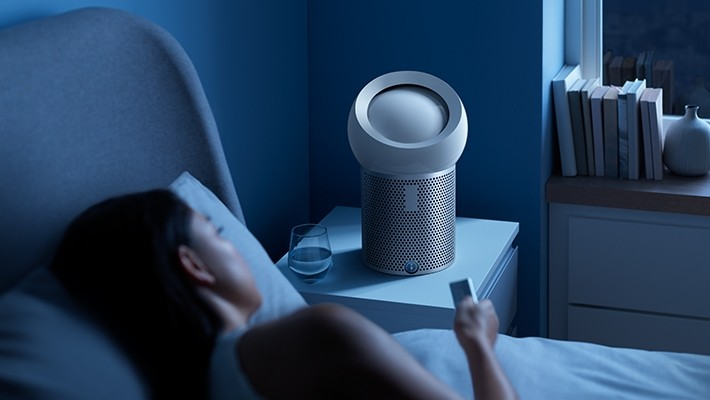 Eine Person im Bett mit einem Ventilator auf dem Nachttisch, einem Glas Wasser und einem Fenster im Hintergrund.
