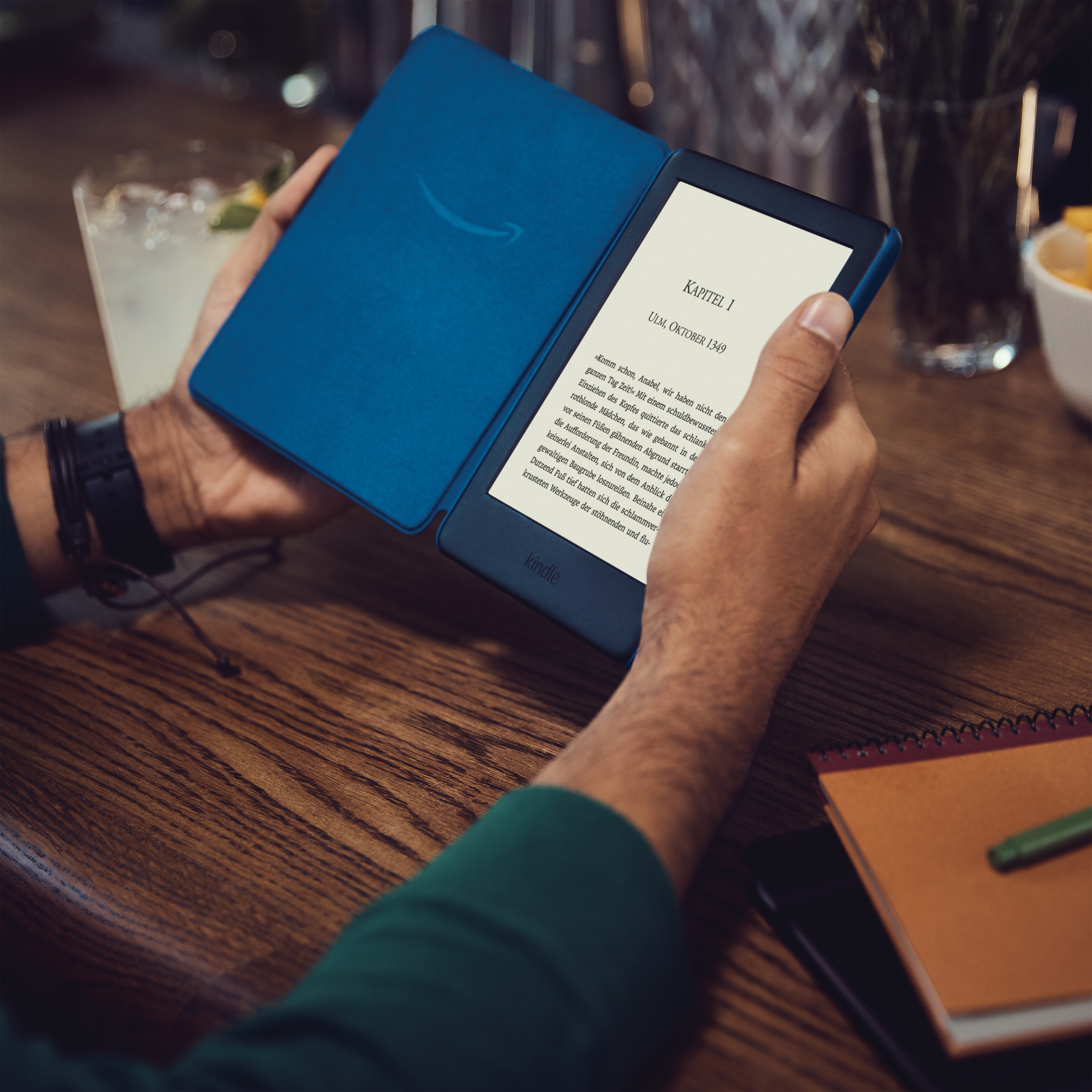 Person hält geöffnetes blaues Kindle mit Text, auf Holztisch mit Getränk.