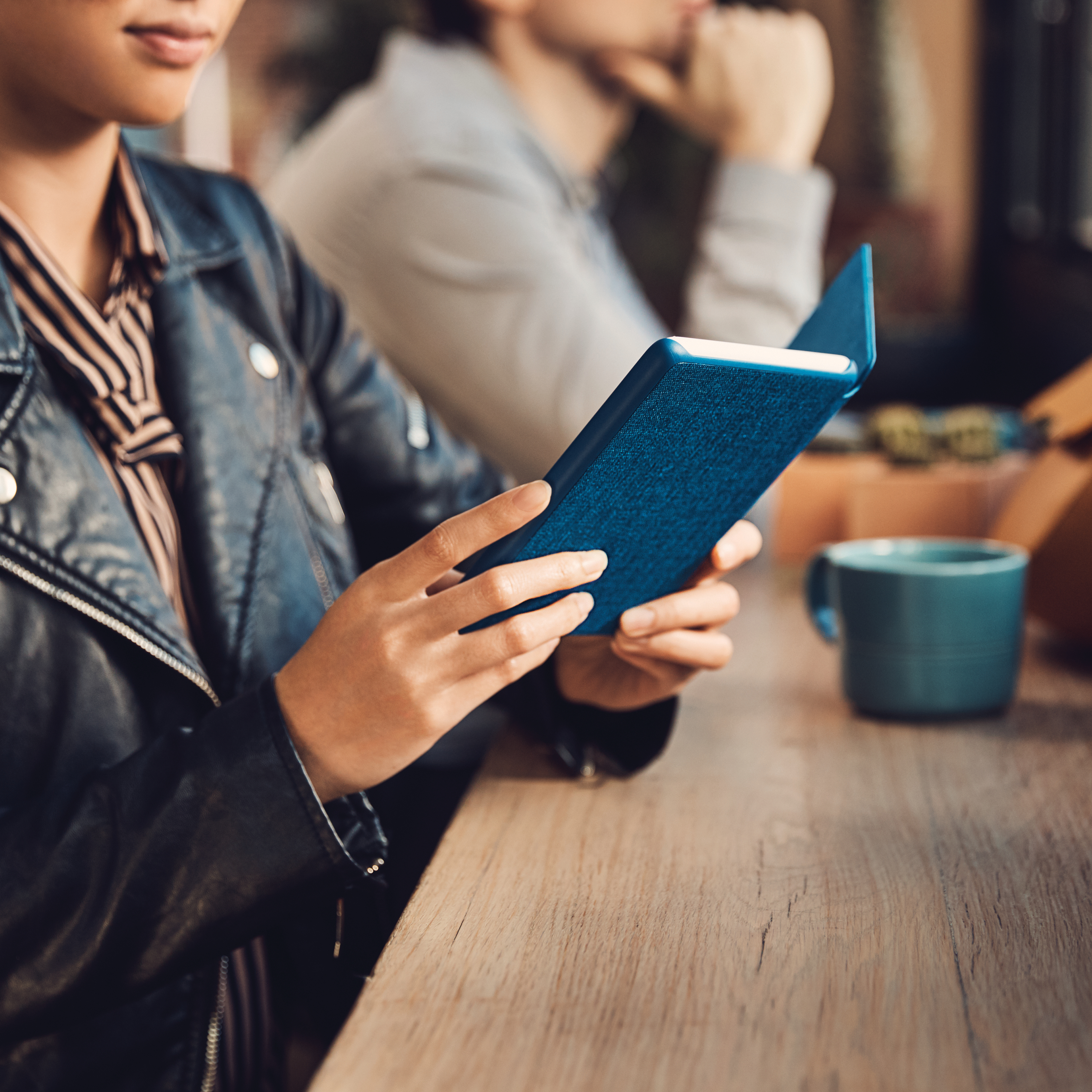 Person hält blaues Buch, sitzt an Holztisch, andere Person dabei.