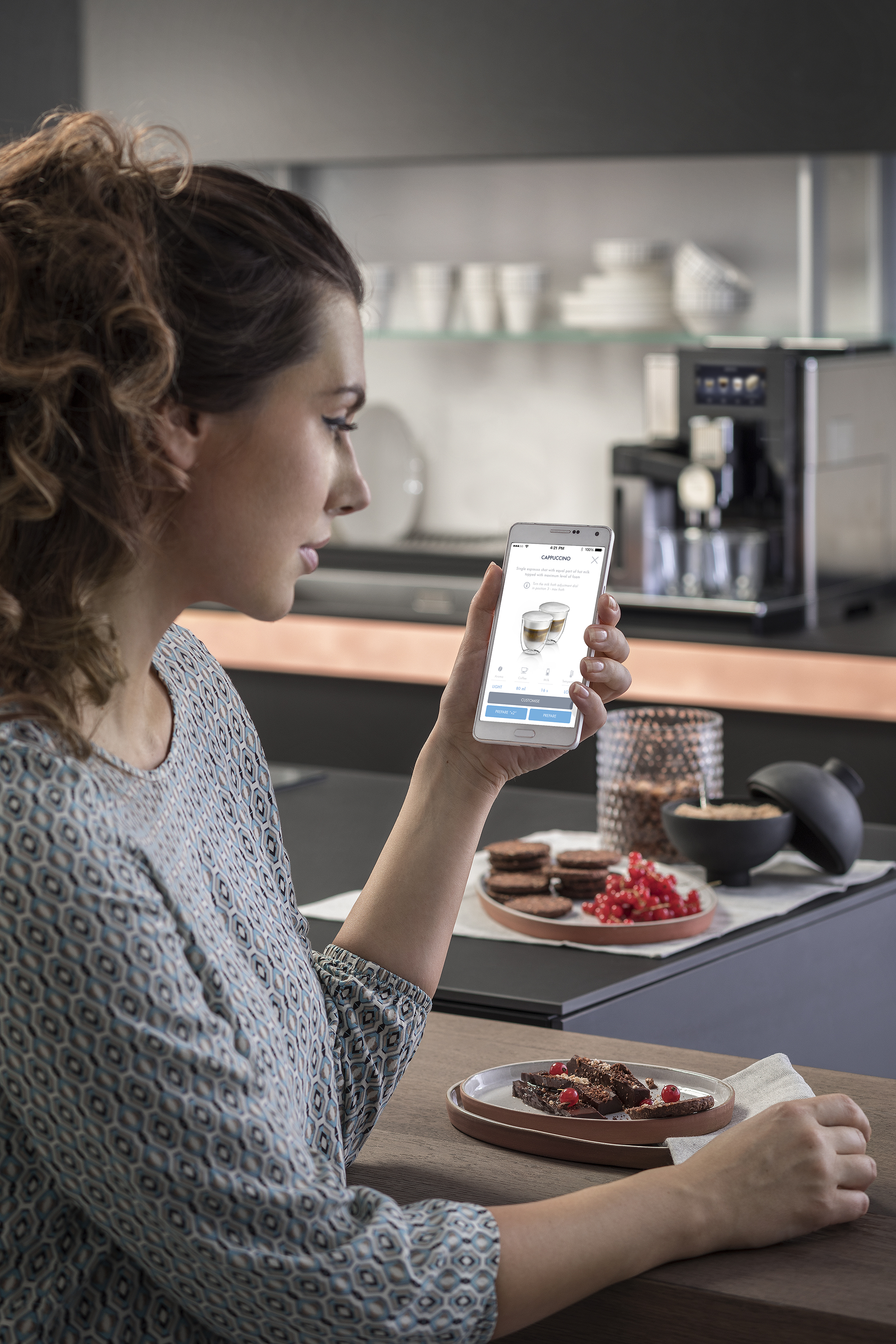 Frau benutzt Smartphone mit Kaffeemaschinen-App, Keksen und Brownie.
