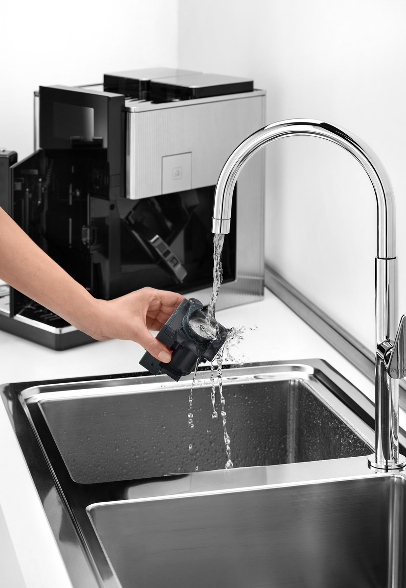 Person wäscht ein schwarzes Kaffeemaschinenteil unter fließendem Wasser in einem Edelstahlbecken.