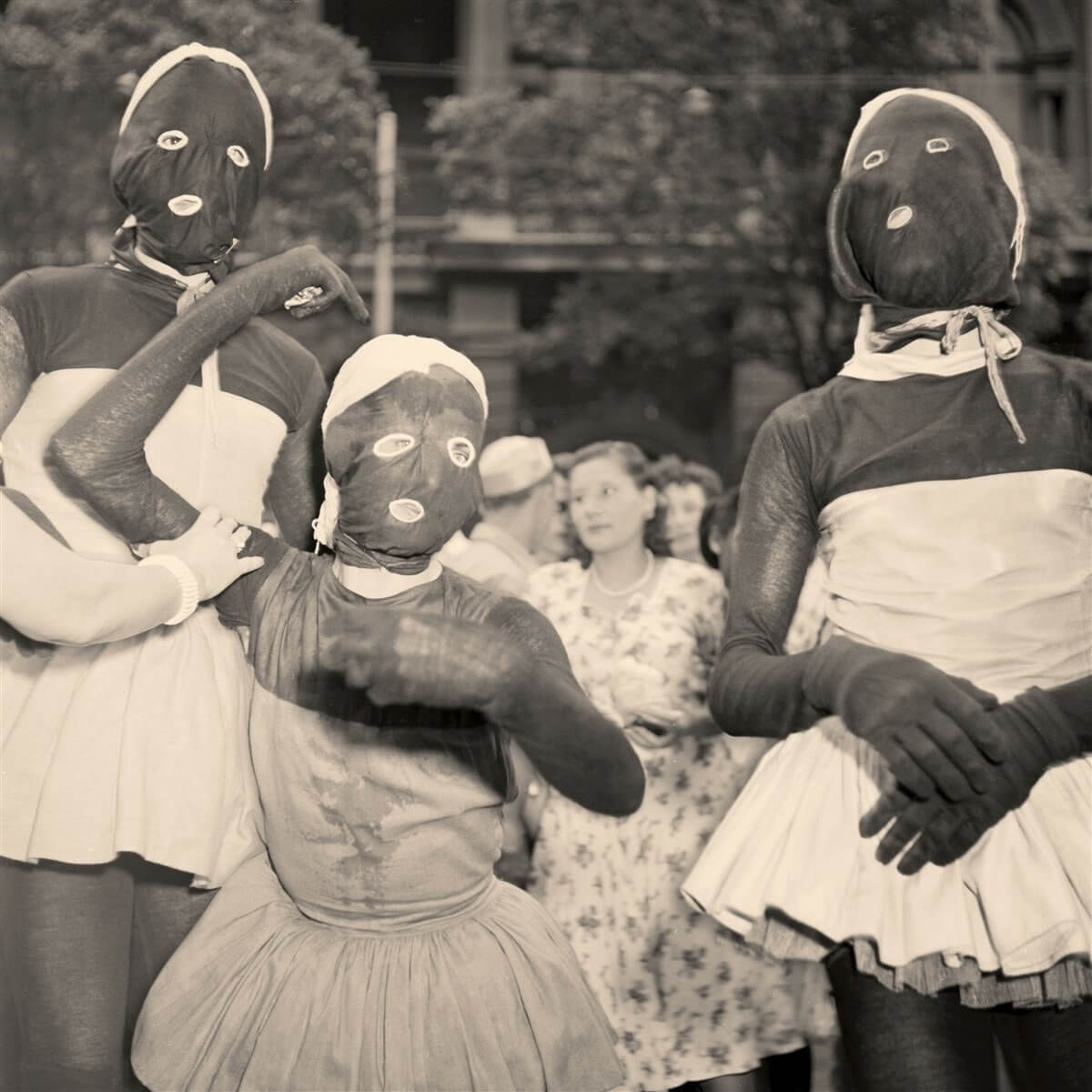 Groupe de personnes en noir et blanc, certaines portant des masques. Le fond est flou. La scène semble vintage.