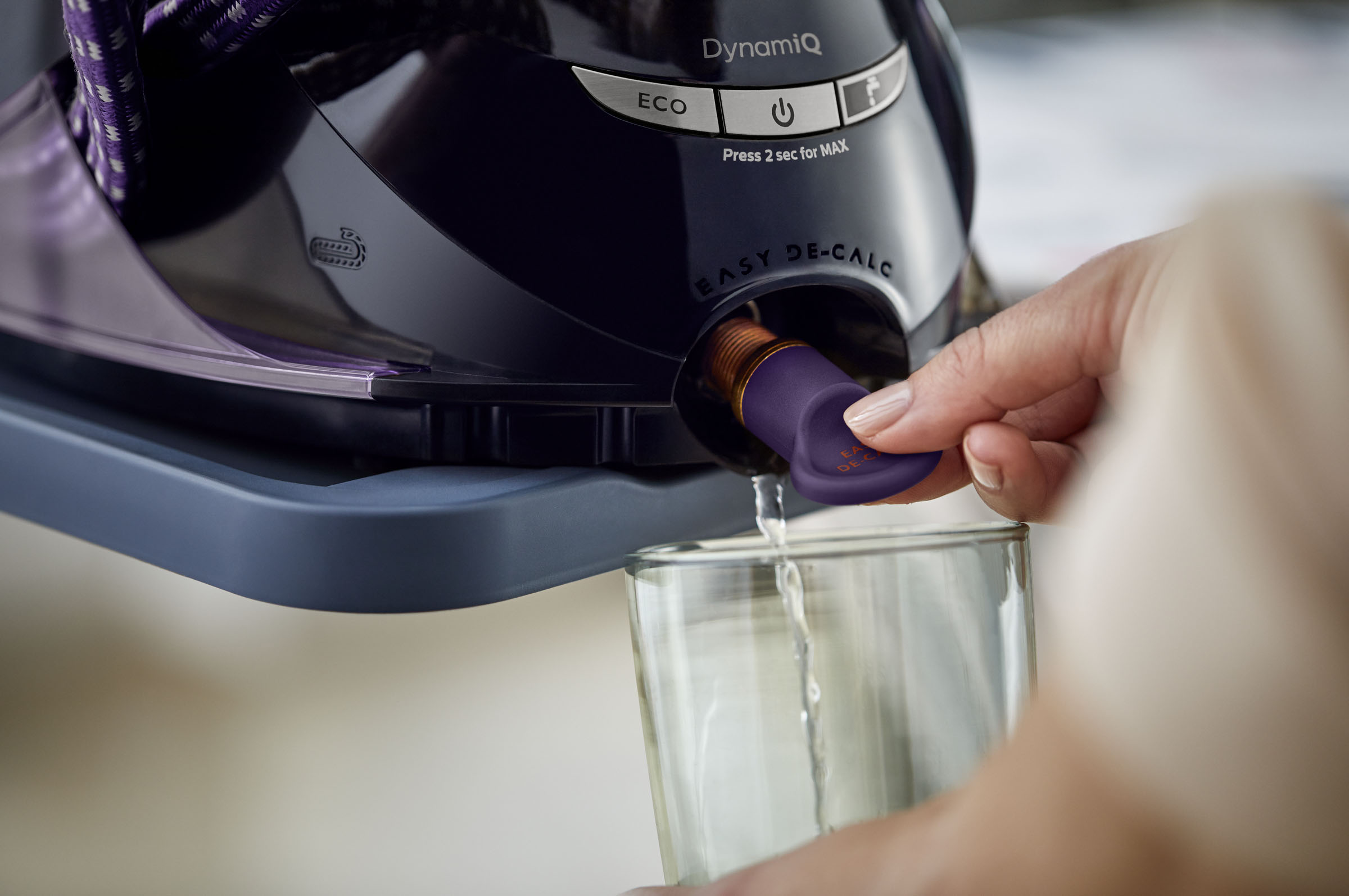 Nahaufnahme einer Hand, die ein Glas mit Wasser aus einem lila-schwarzen Bügeleisen füllt. Dampf kommt heraus.