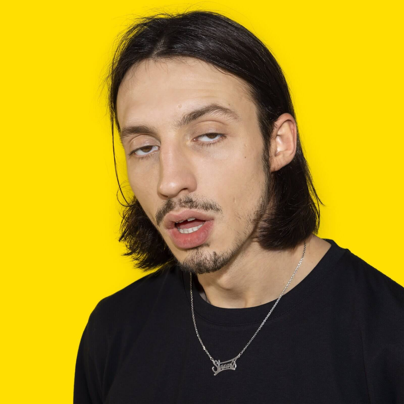 Een jonge man met lang donker haar en een sik, draagt een zwart shirt, gele achtergrond.