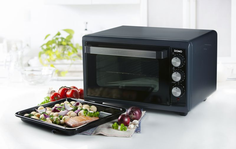 An oven with vegetables and a tray of food, on a white countertop.