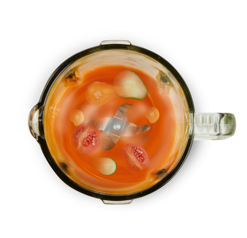 Top view of a blender with orange liquid, and cut tomatoes and cucumbers.