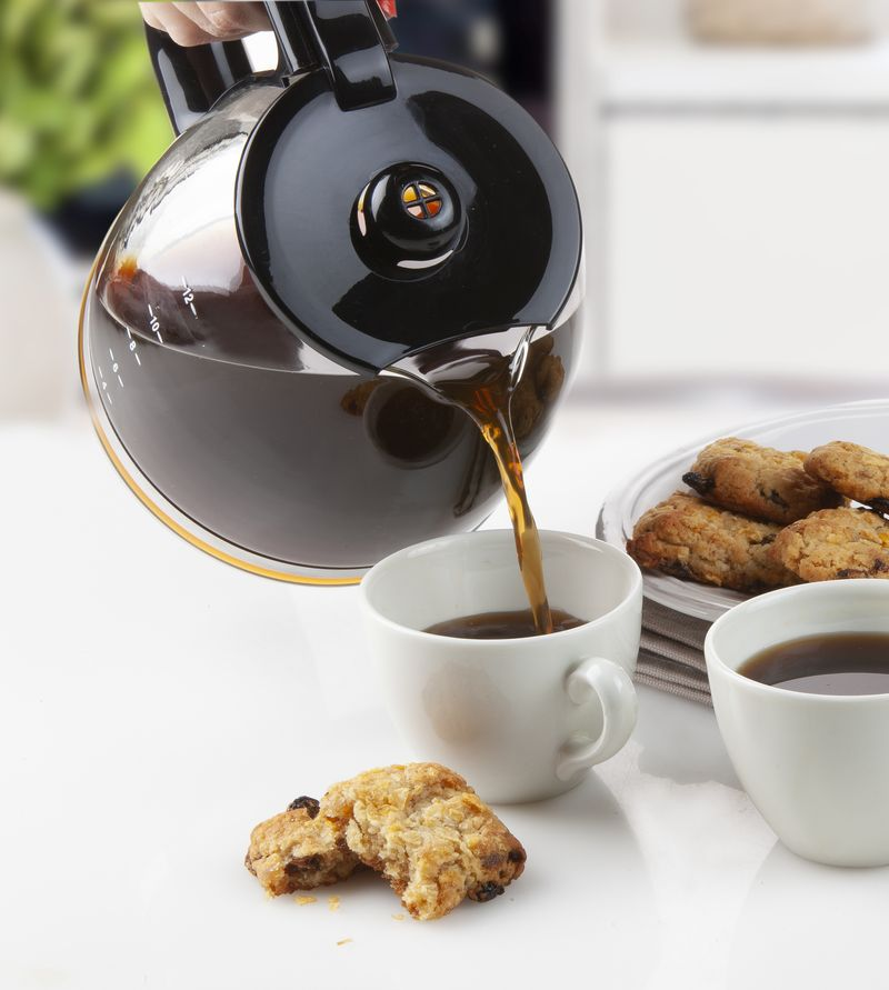 Coffee pours from a glass carafe into a white cup, with cookies nearby on a white surface.