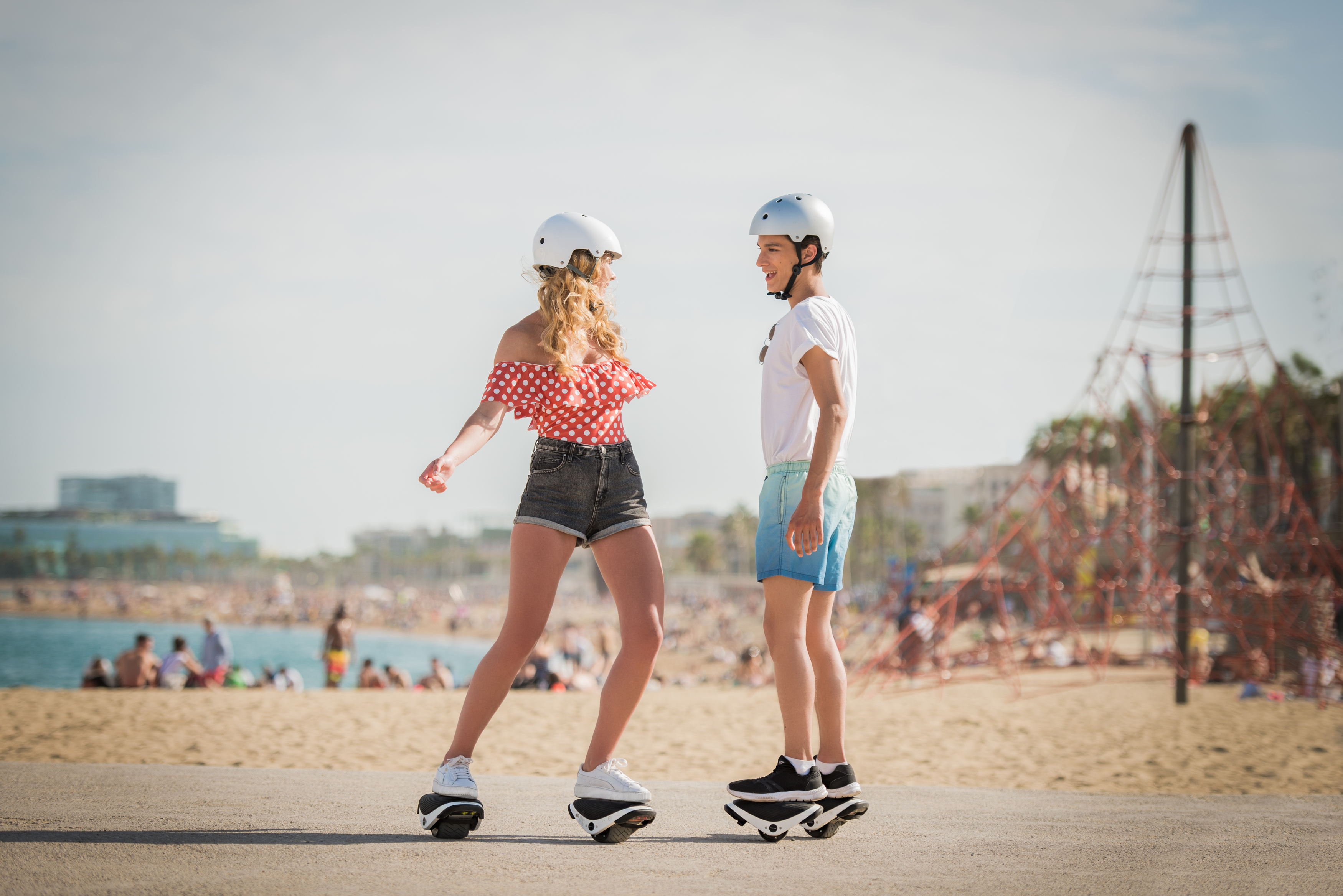 Zwei Personen auf Hoverboards auf einer gepflasterten Oberfläche, mit Strand und Meer im Hintergrund.