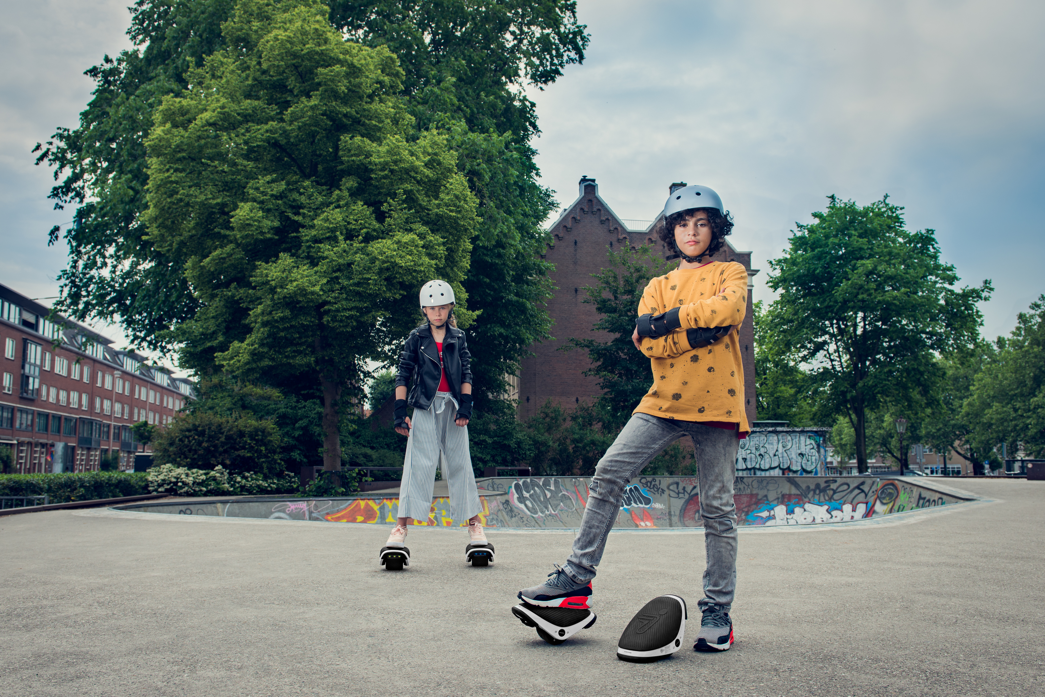 Zwei Kinder auf Hover-Schuhen in einem Skatepark. Bäume und Gebäude im Hintergrund.