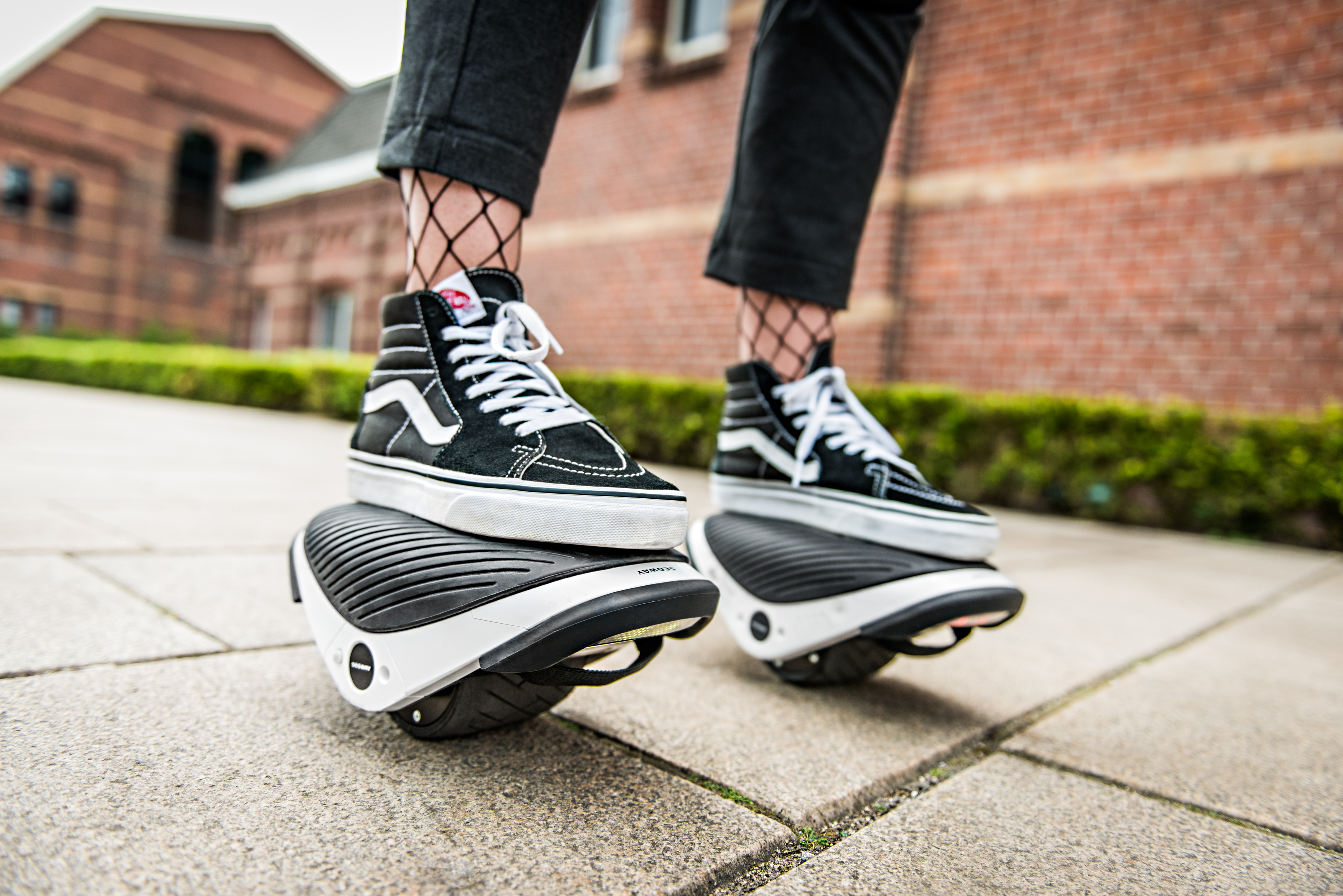 Person trägt schwarz-weiße Sneaker auf Segway-ähnlichen Geräten. Der Hintergrund ist verschwommen.