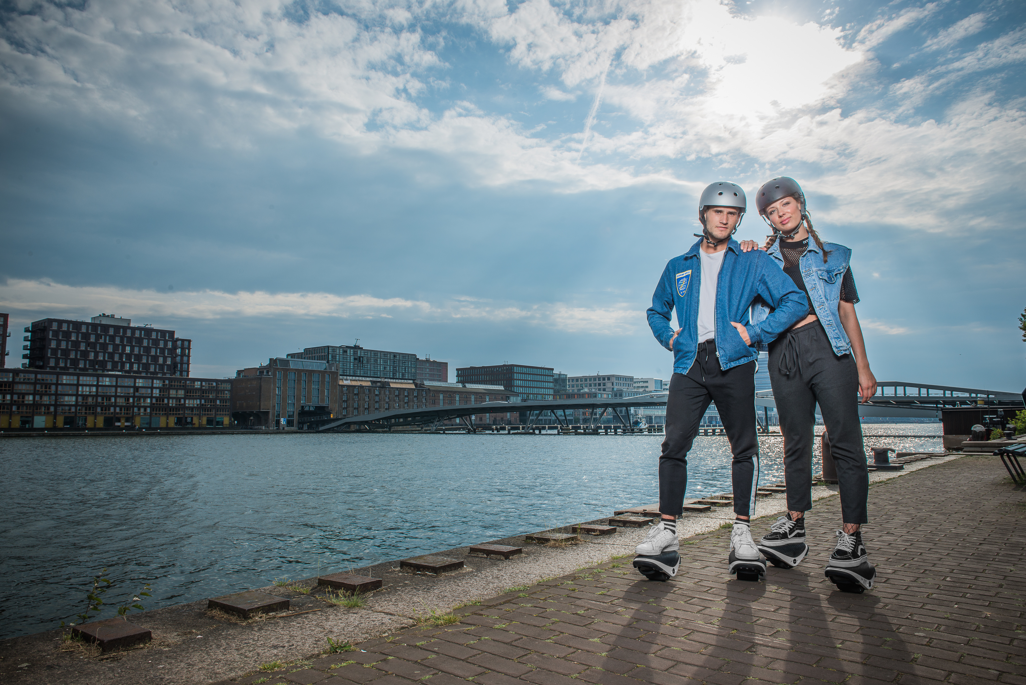Zwei Personen auf Hoverboards in der Nähe eines Gewässers. Wolkenverhangener Himmel mit Gebäuden im Hintergrund.