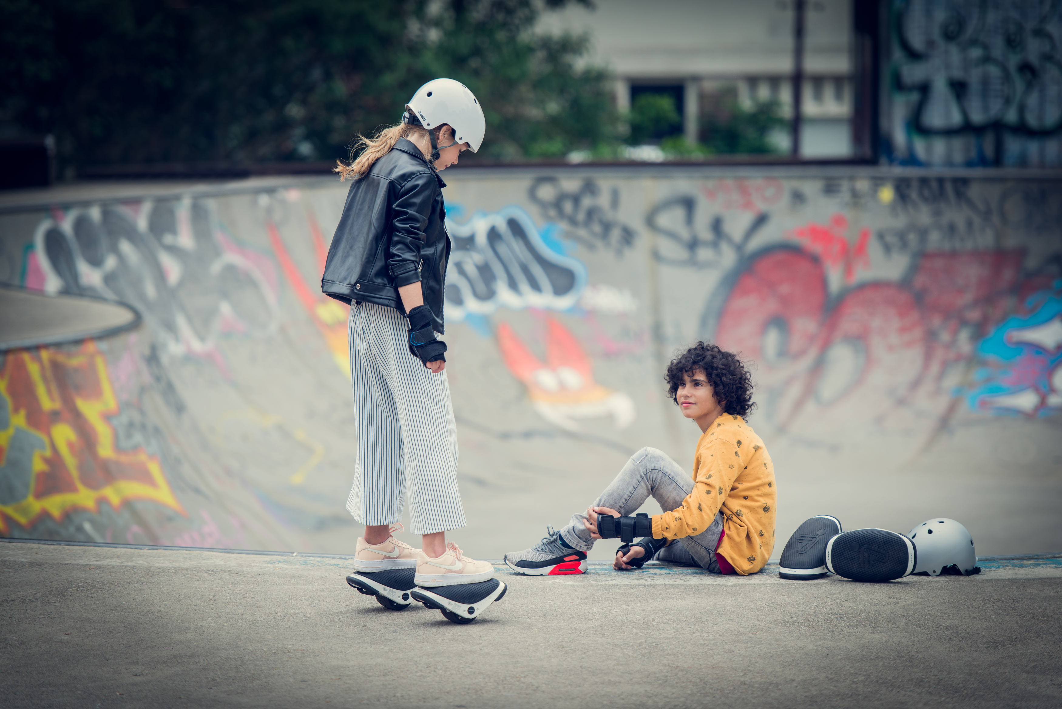 Zwei Kinder in einem Skatepark. Eines auf Hover-Schuhen, das andere sitzt. Betonbecken im Hintergrund.