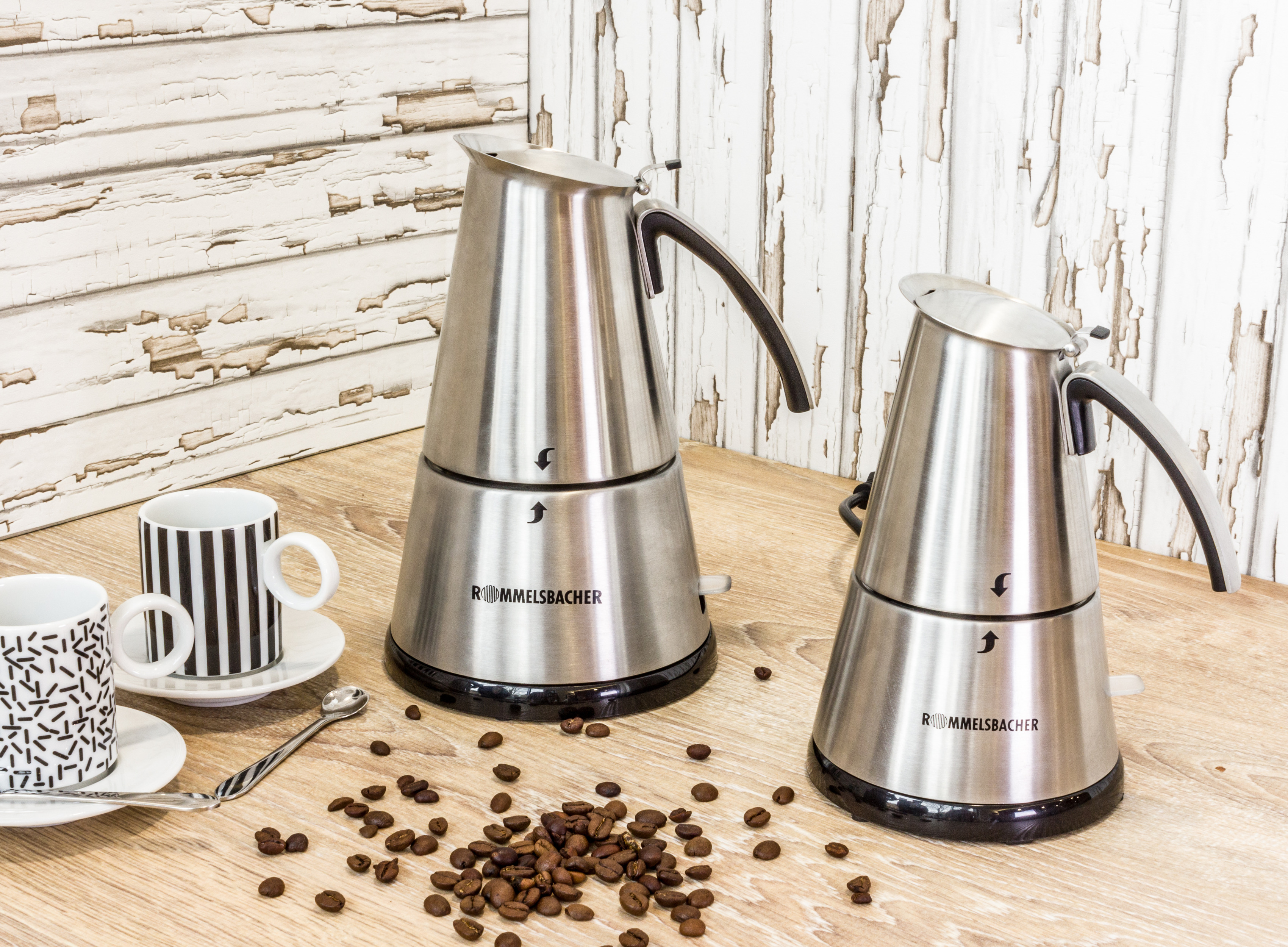 Zwei Kaffeemaschinen aus Edelstahl, zwei Tassen, ein Löffel und Kaffeebohnen auf einer Holzoberfläche mit weißem Hintergrund.