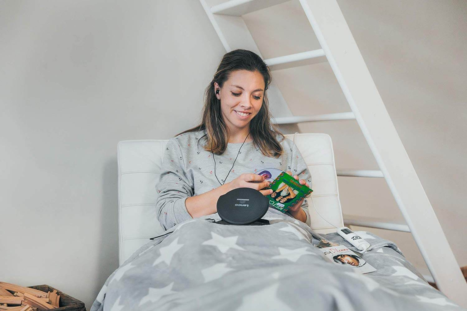 Woman in grey top listening to music from a black CD player, sitting with a blanket.