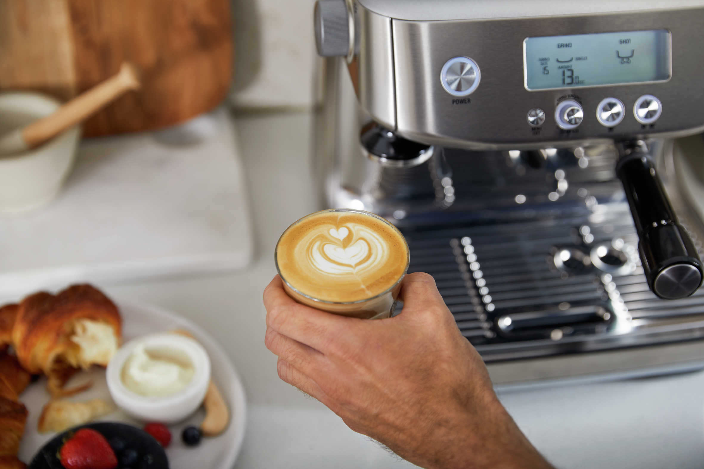 Eine Hand hält einen Latte mit Herzdesign, in der Nähe einer Kaffeemaschine und Gebäck.