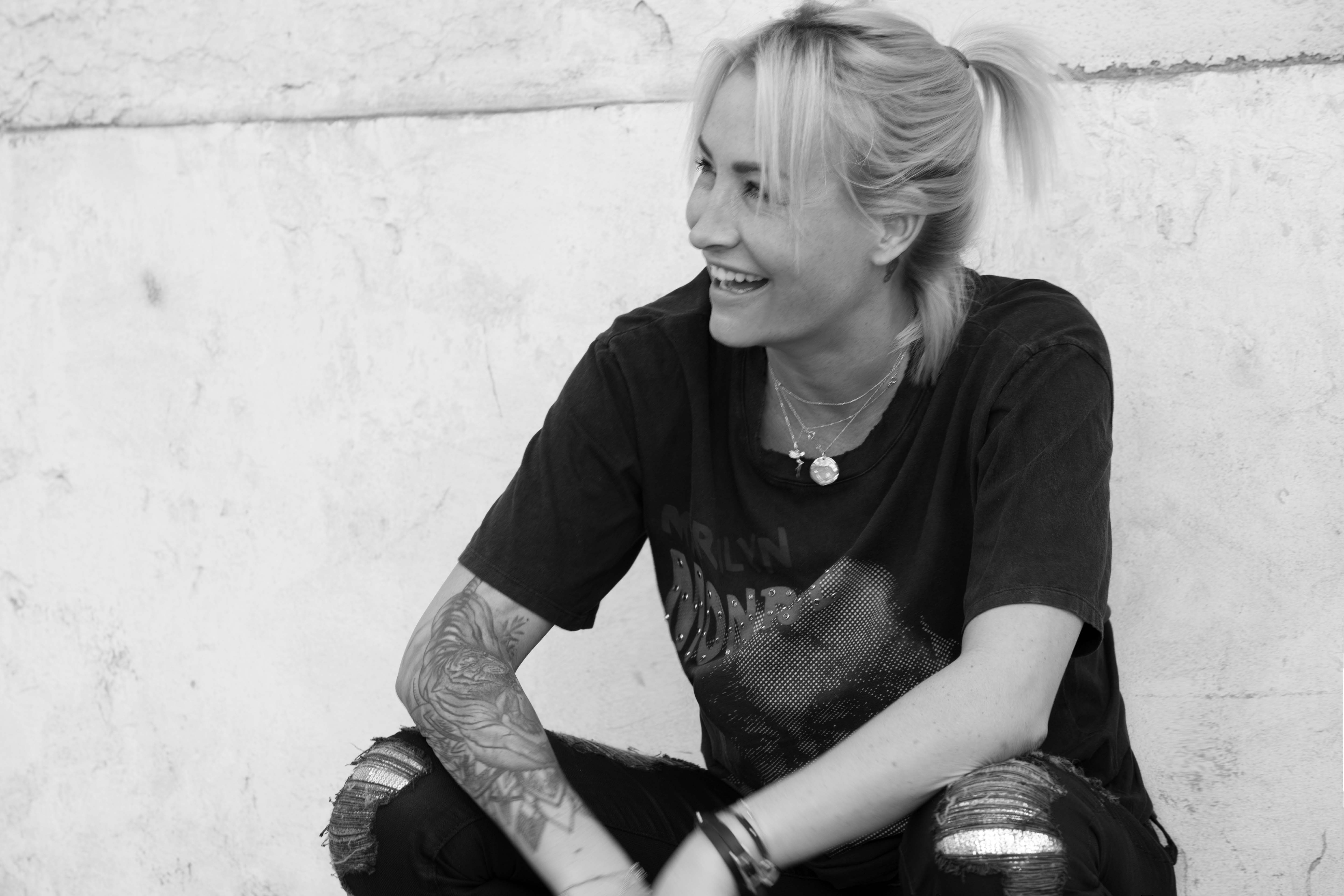 Blonde Frau lächelt und schaut nach rechts. Sie trägt ein schwarzes Hemd und hat Armtattoos. Weiße Wand.