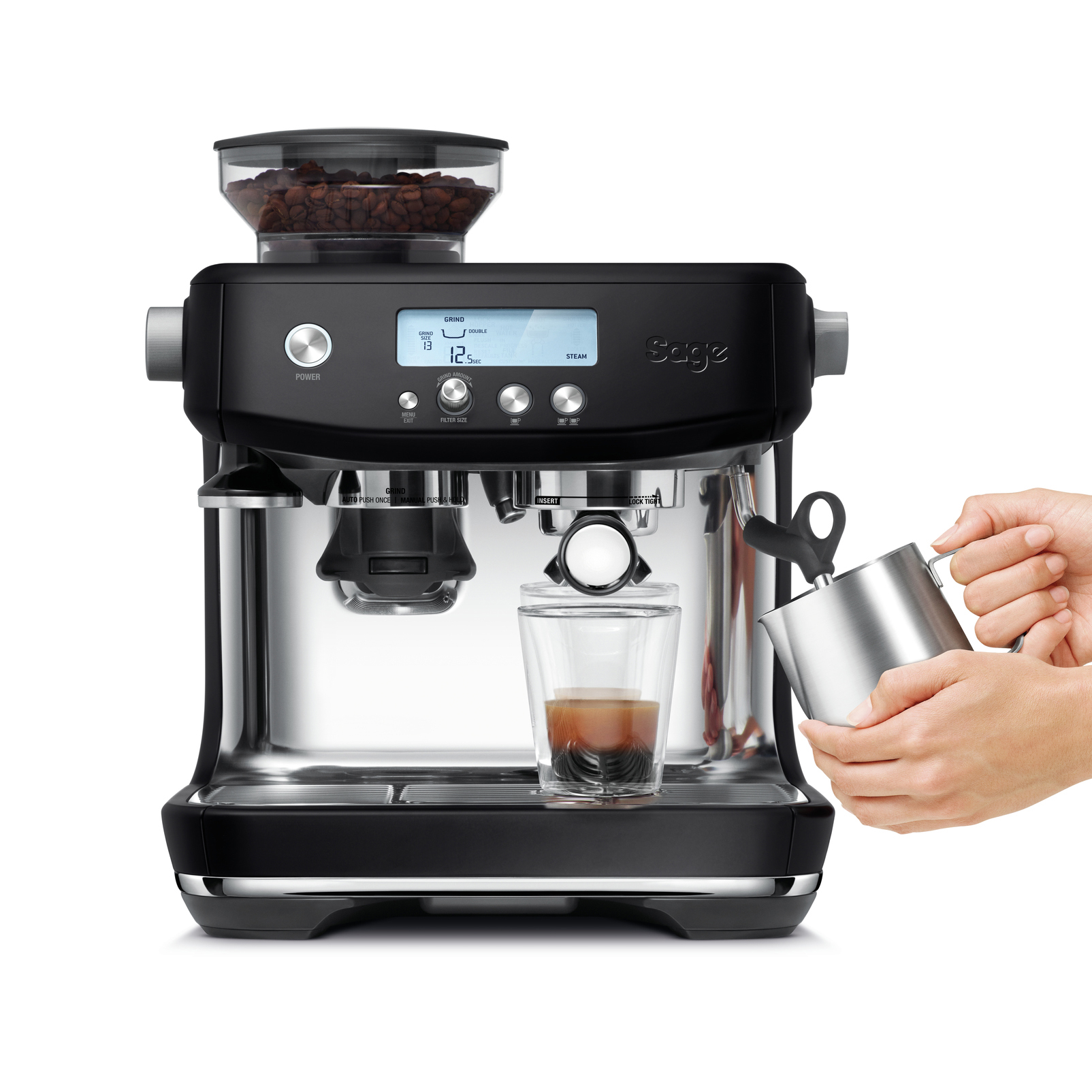 Schwarze Espressomaschine. Eine Hand gießt Milch ein. Ein Glas enthält Espresso. Kaffeebohnen oben.