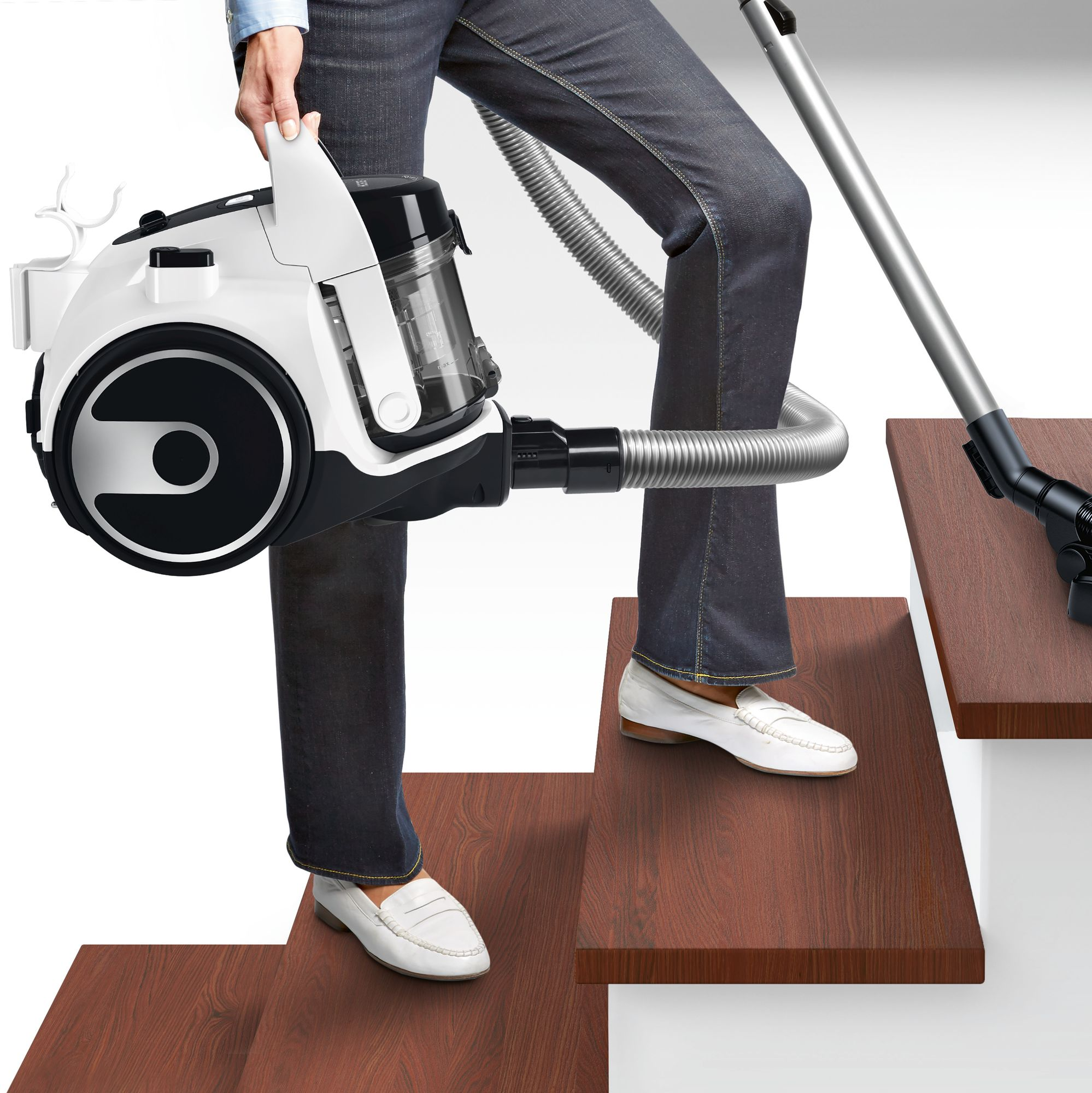 A person holding a white and black vacuum cleaner, vacuuming stairs. They wear jeans and white shoes.