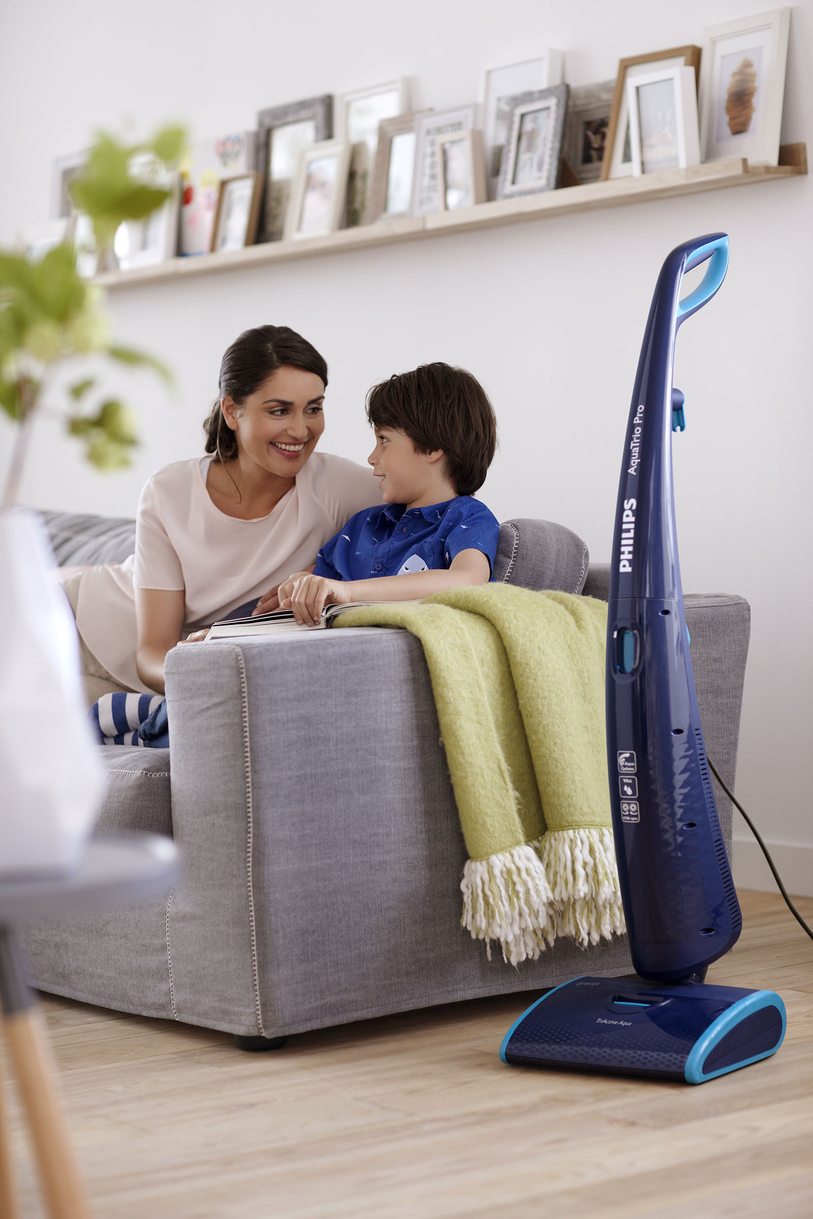 Frau und Kind auf dem Sofa, schauen sich an. Staubsauger im Hintergrund. Eine Decke liegt auf dem Sofa.