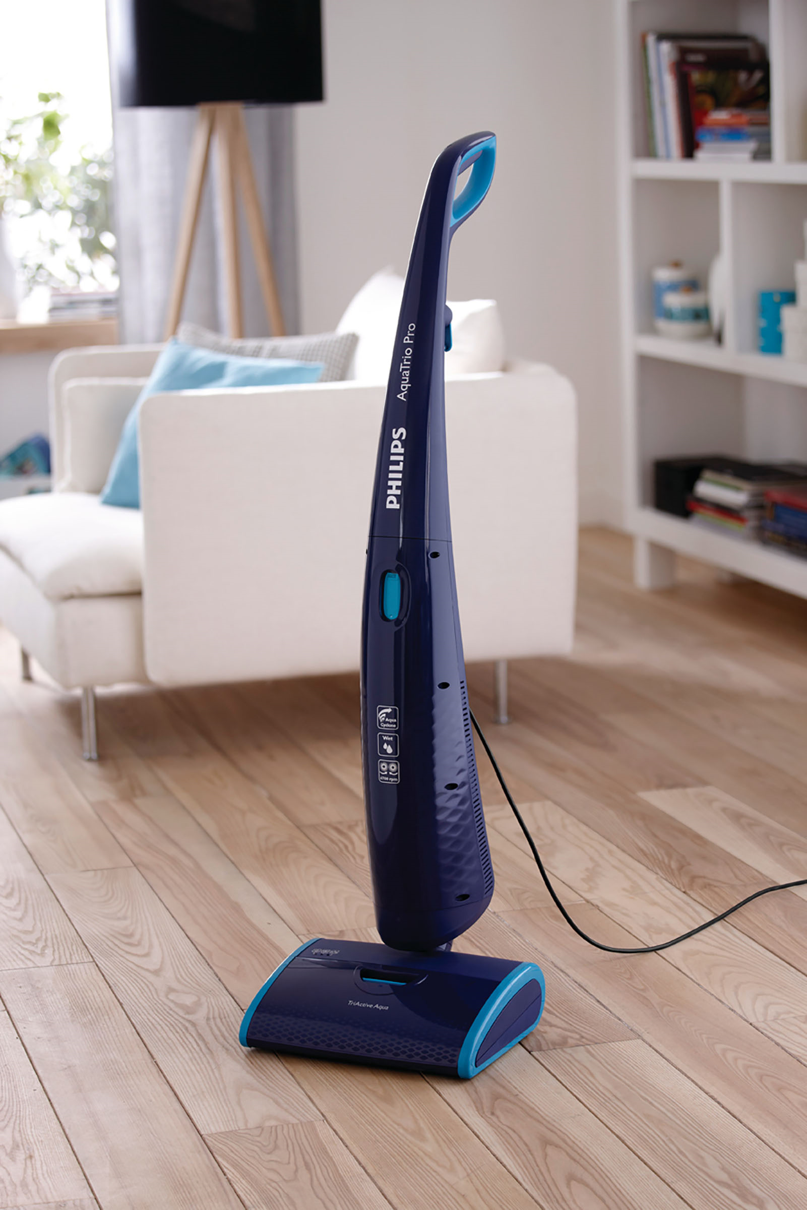 Ein blauer Philips AquaTrio Pro Staubsauger steht auf einem Holzboden. Im Hintergrund sind ein weißes Sofa und ein Bücherregal.