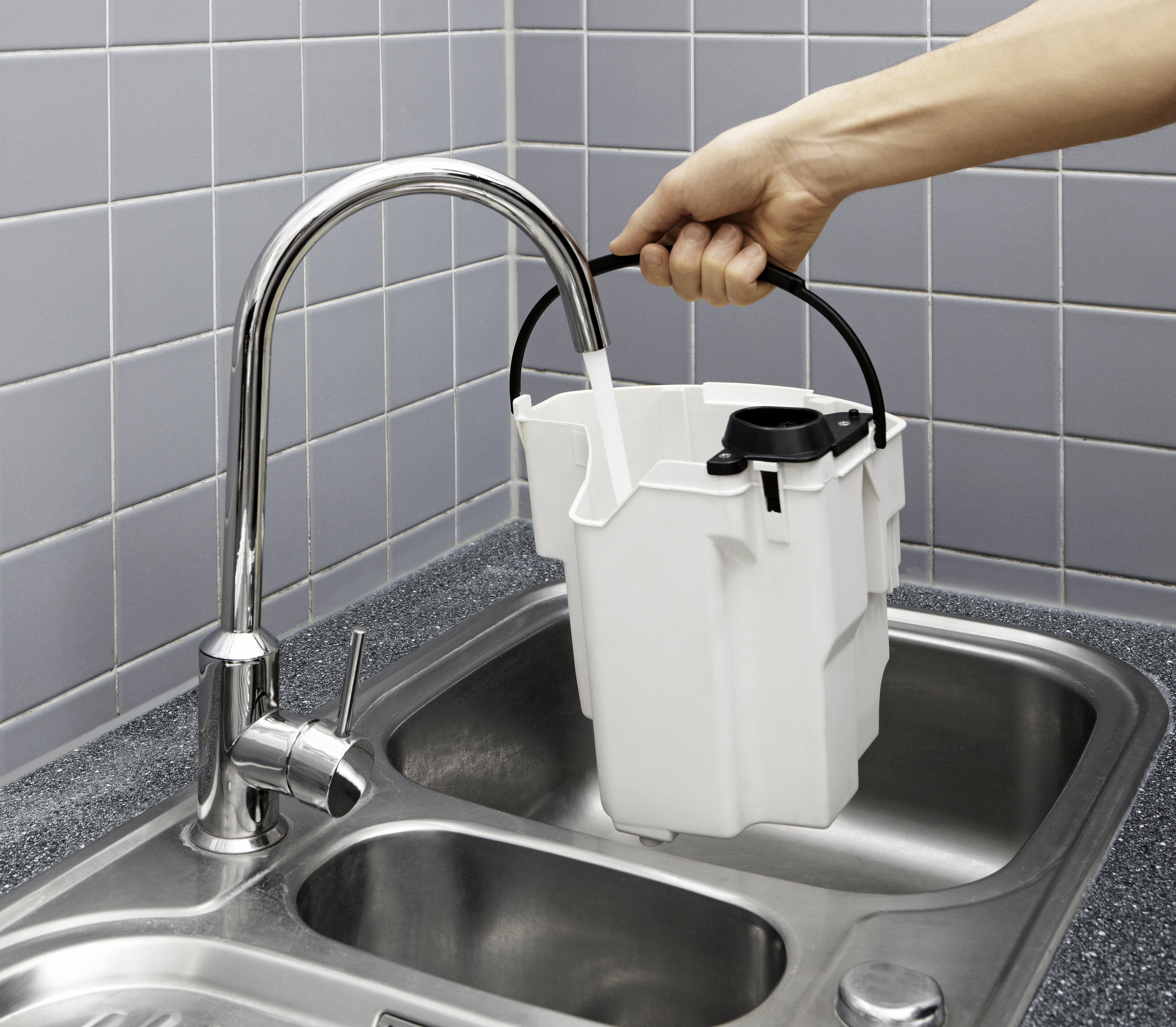 Eine Person, die einen weißen Eimer unter einem Chromhahn hält und ihn in einem Küchenbecken mit Wasser füllt.