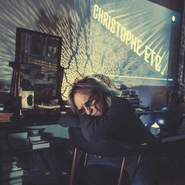 Homme avec des lunettes de soleil appuyé sur un bureau, un écran affiche 'CHRISTOPHE ETC'. Scène sombre.
