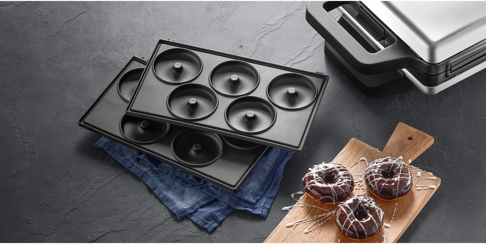 Overhead shot: donut maker, donuts on a wooden board, and a waffle maker. The background is a dark grey surface with a blue cloth.