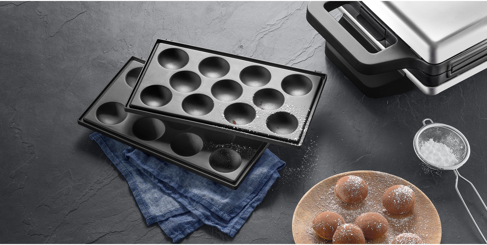 Black takoyaki maker, powdered sugar sifter, plate of balls, and waffle maker on dark surface.